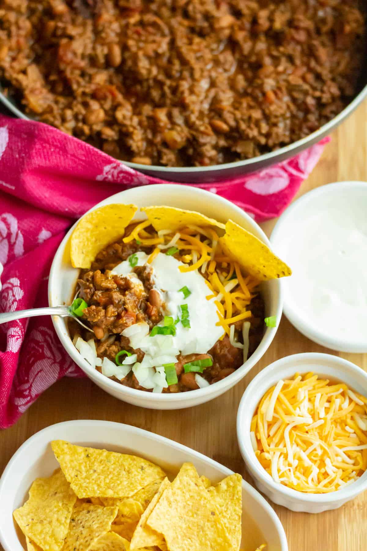 Overhead shot of bowl of chili  with chips, sour cream, cheese and green onions and condiments in the background