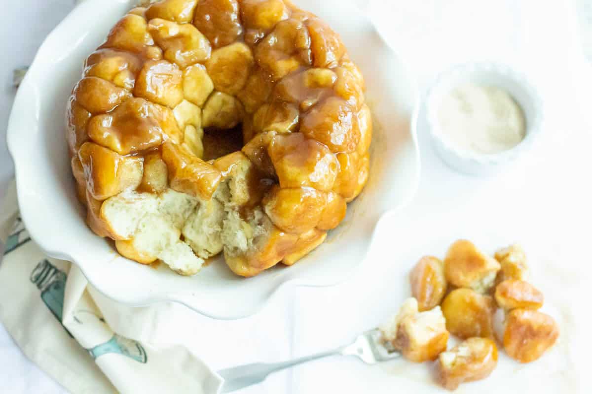 Overhead shot of monkey bread on a white cake plate with a small bowl of sugar in the back and a plate of monkey bread with a fork in the front