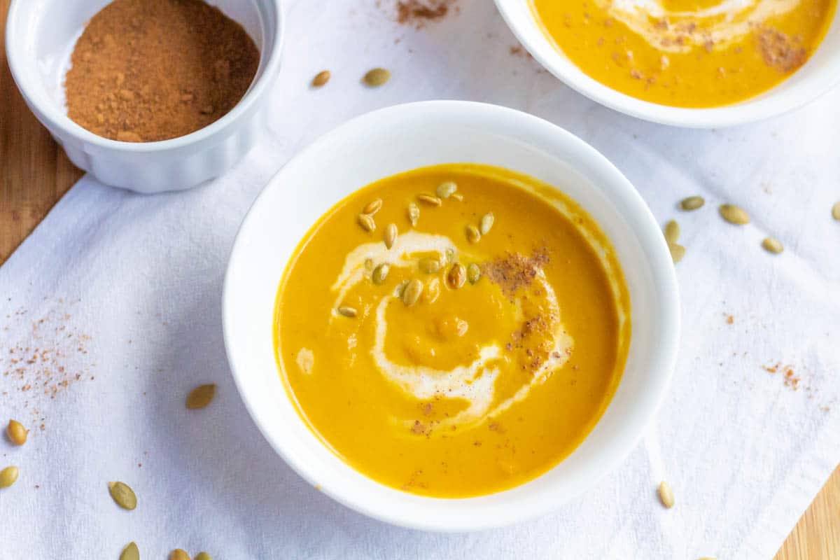 Two bowls of pumpkin bisque with a ramekin with nutmeg in the background