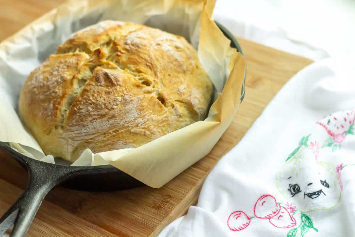 Crusty No Knead Bread | thewendthousekitchen.com Baked bread in a cast iron pan with parchment paper and a tea towel next to it