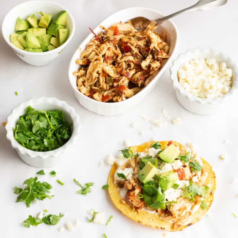 Tinga on a tostada with more chicken, cheese, cilantro and avocado in the background in bowls