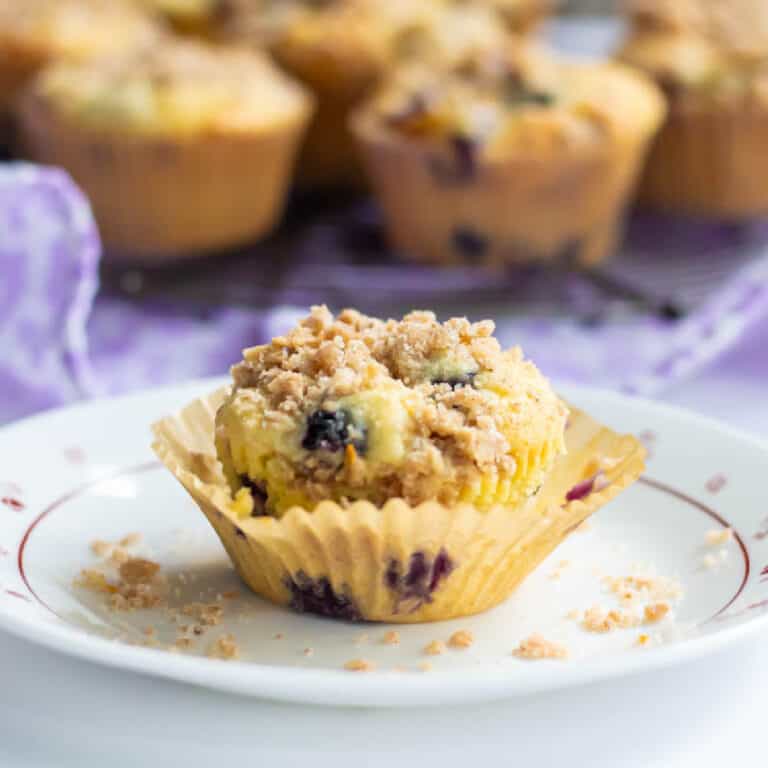 A blueberry muffin inside an open cupcake liner with more muffins in the background