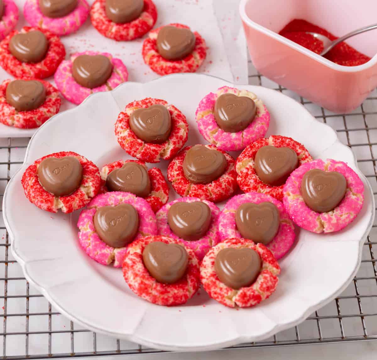 A plate of Valentine sugar cookies with other cookies in the background and a small pink bowl of sanding sugar