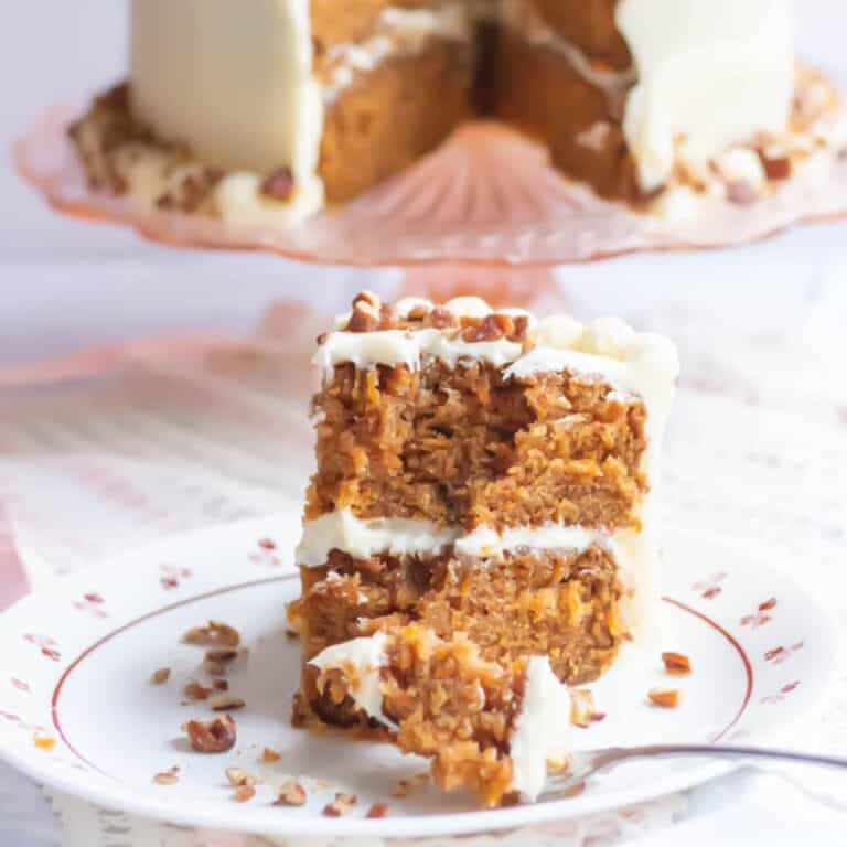 Carrot cake on a white plate with a fork and the whole carrot cake in the background on a pink cake plate