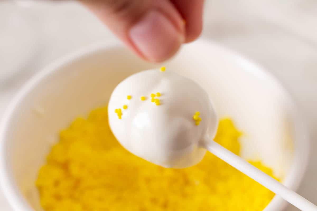 Holding a cake pop over yellow nonpareils and adding some to the cake pop