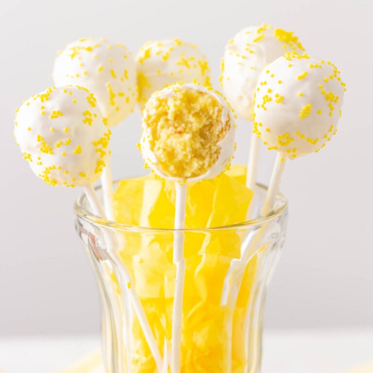 Cake pops in a tall glass with yellow tissue paper and the cake pop in the front has a bite taken out of it