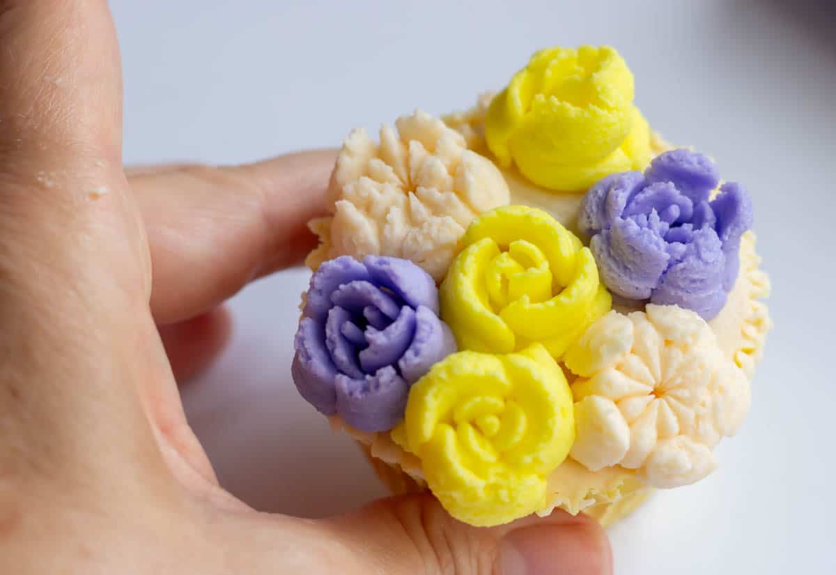 Buttercream flowers in lavender, yellow and ivory covering an iced cupcake