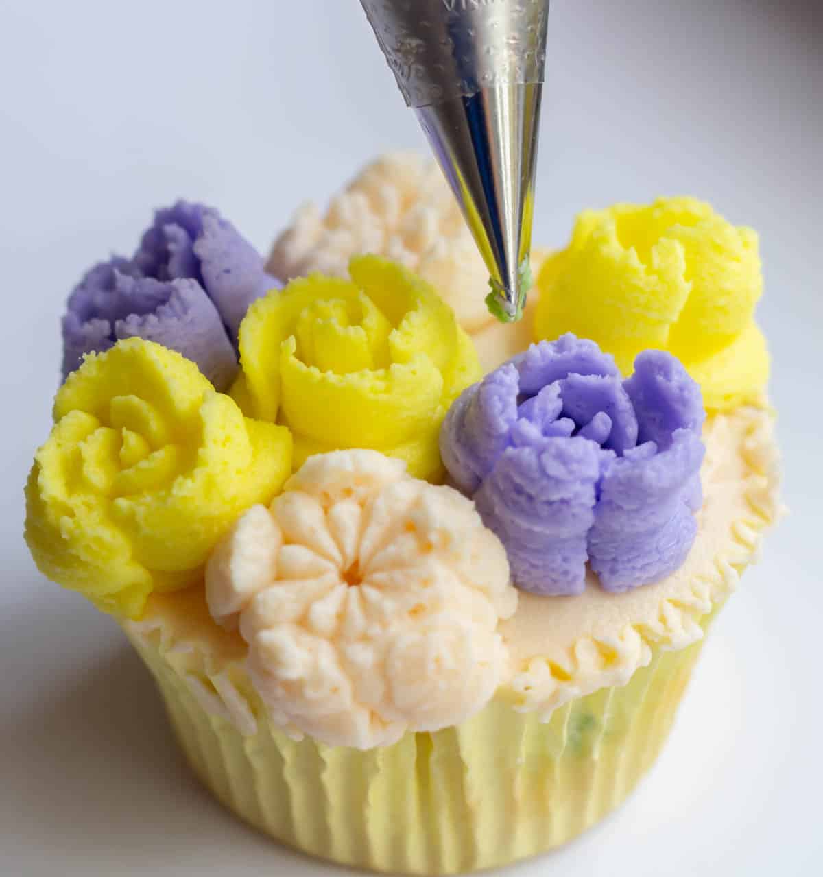 Holding a piping bag fitted with green buttercream and a #352 leaf tip in between buttercream flowers on a cupcake