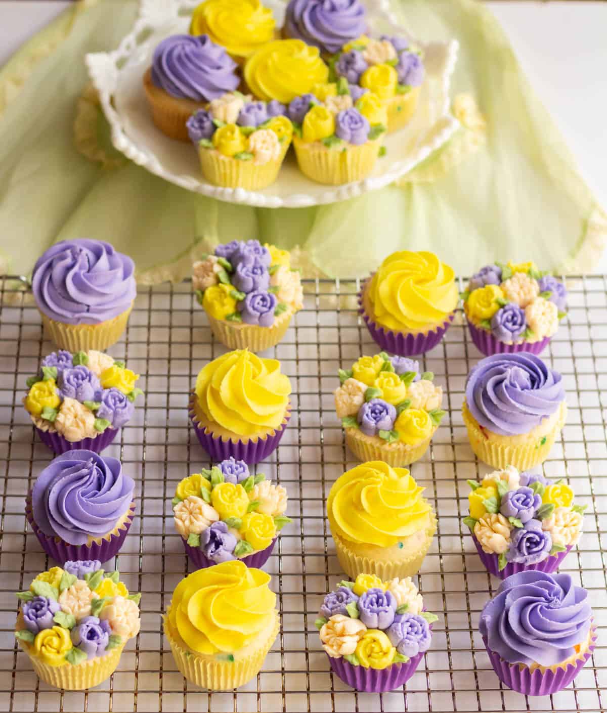Decorated lavender and yellow cupcakes on a cooling grid with decorated cupcakes in the background on a white cake pedestal