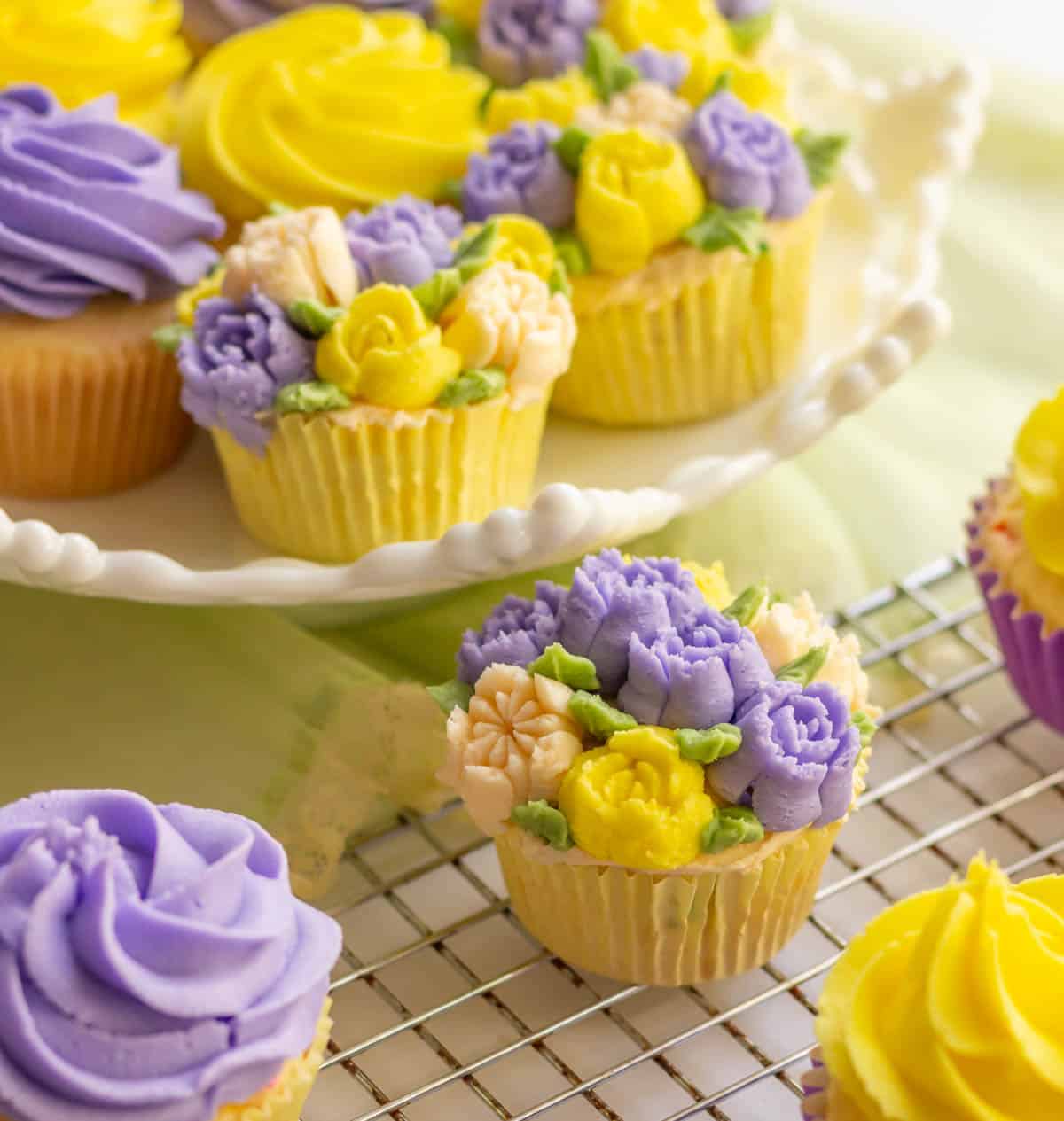 Decorated lavender and yellow cupcakes on a white cake stand and some in the front on a wire grid