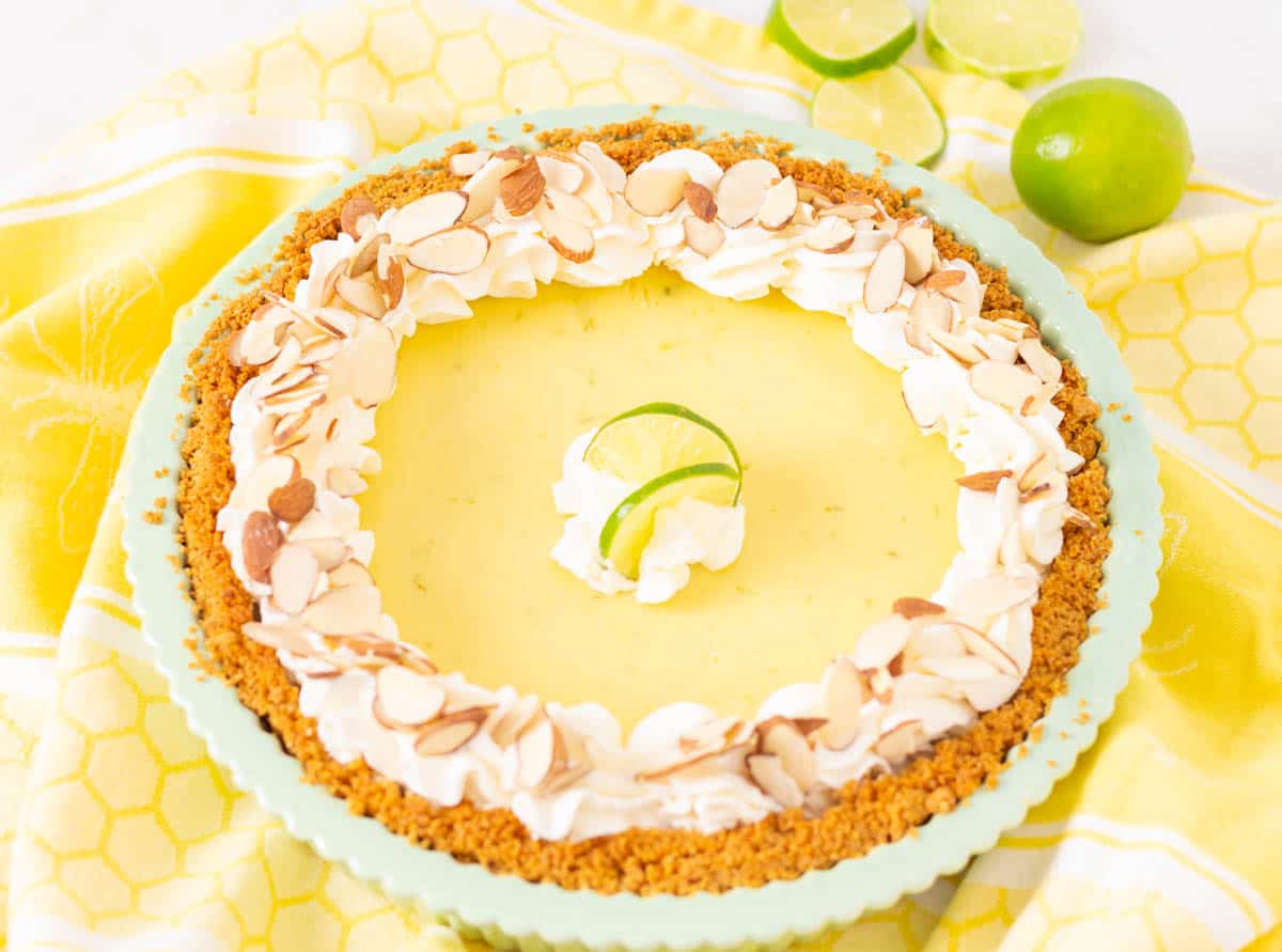 Key lime pie with whipped cream and almonds in a green pie plate with limes in the background