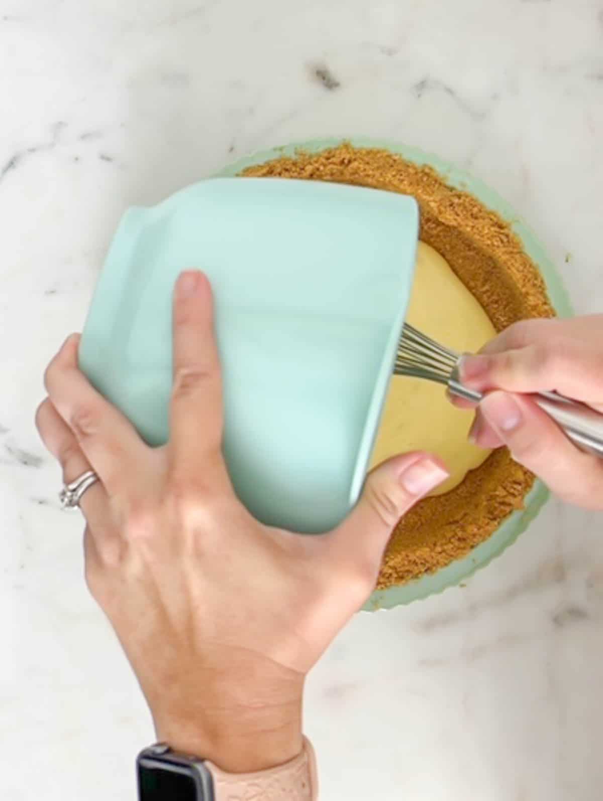 Pouring key lime pie filling into graham cracker crumbs in a green pie plate