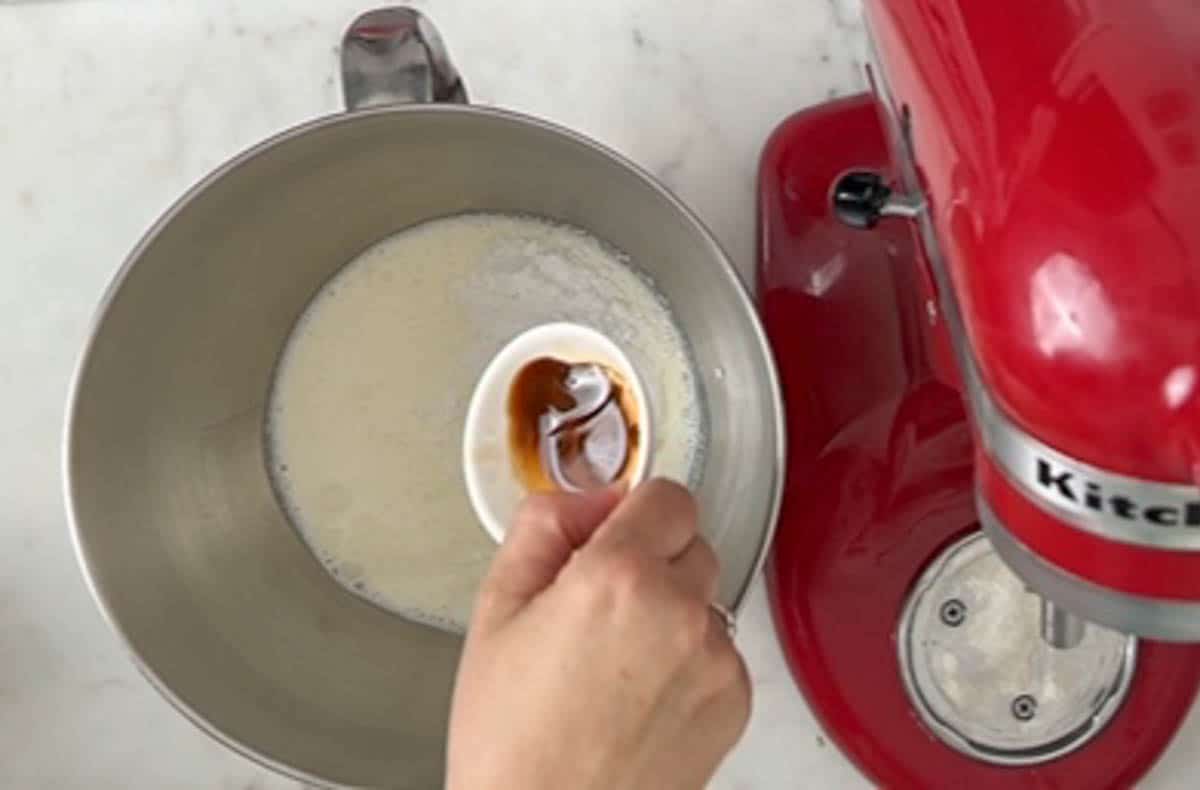 Pouring vanilla extract into a bowl of a red stand mixer with heavy cream and powdered sugar