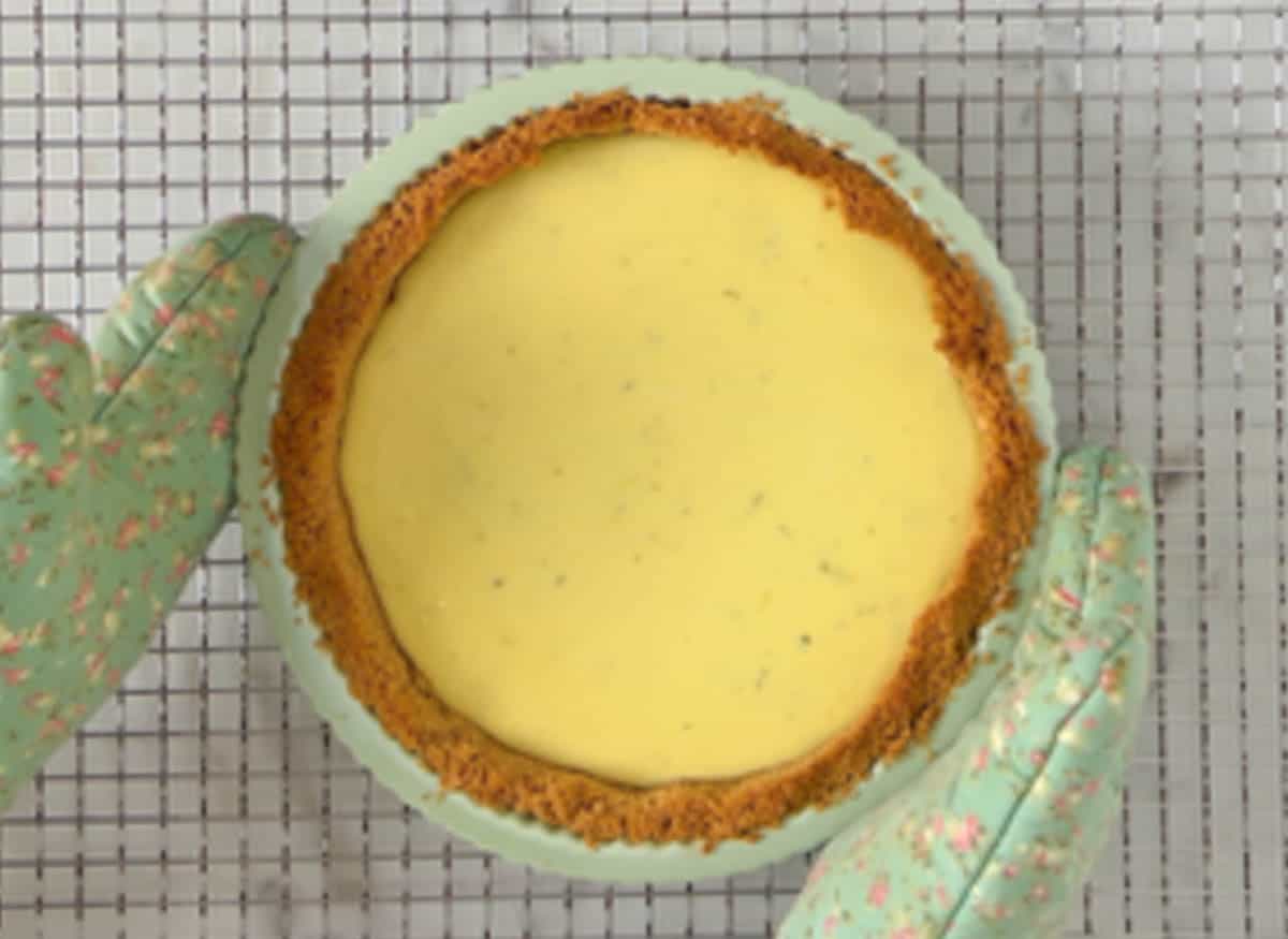 Baked key lime pie on a wire cooling grid being held by two hands with green floral pot holders