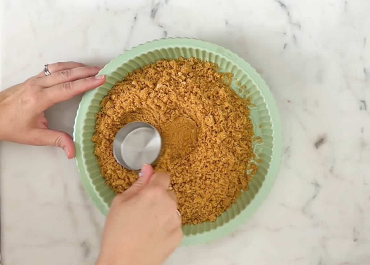 Pressing graham cracker crust into a green pie plate with a metal measuring cup