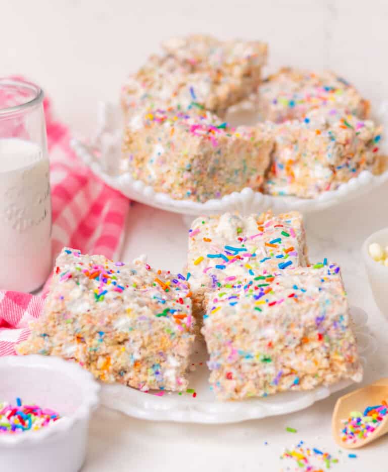 Rice Krispies treats on a white cake pedestal and more on a white plate in front with sprinkles in a bowl