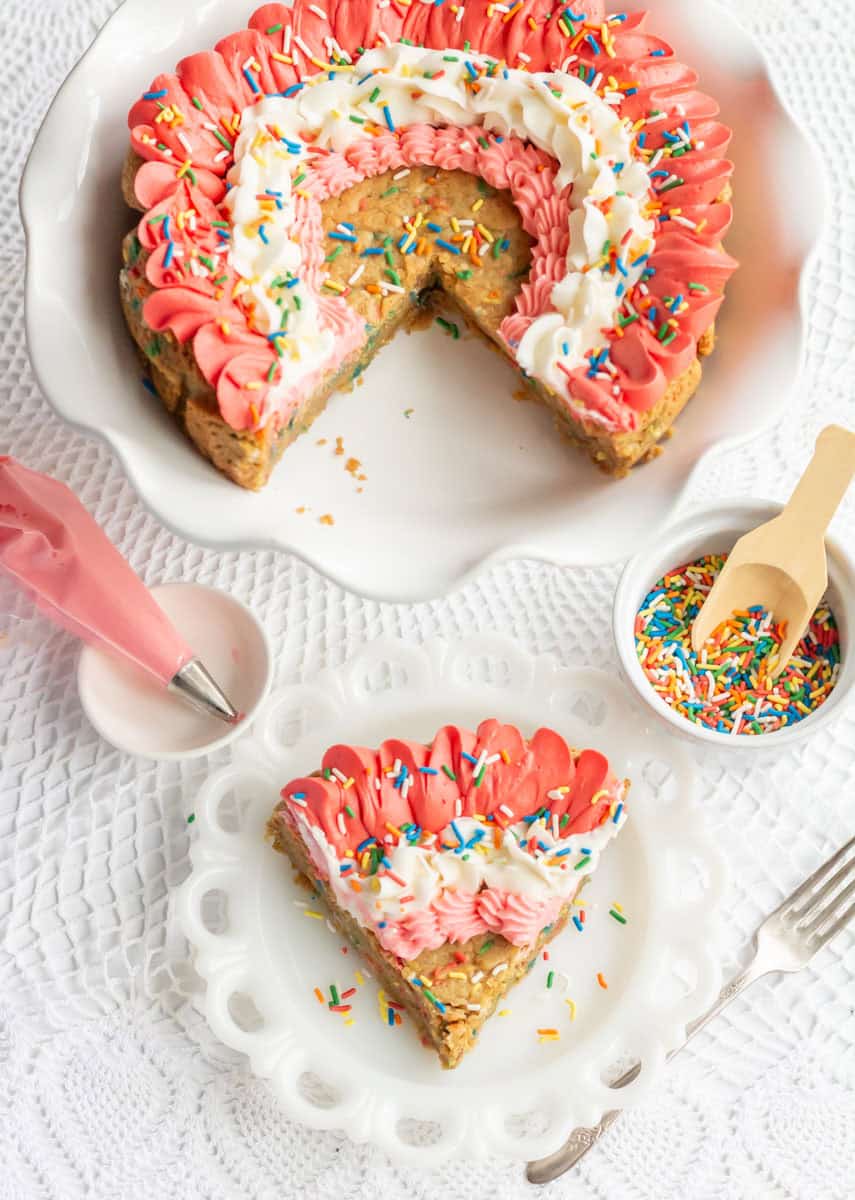 Funfetti Cookie Cake | thewendthousekitchen.com Cookie cake on a with a piece cut out, under that a piping bag with pink frosting and rainbow sprinkles in a bowl. Under that a piece of cookie cake on a white plate.