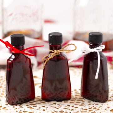 Three bottles of vanilla extract with different color bows tied around the necks. Two mason jars with vanilla extract in the background.