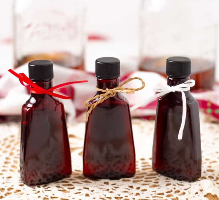 Three bottles of vanilla extract with different color bows tied around the necks. Two mason jars with vanilla extract in the background.