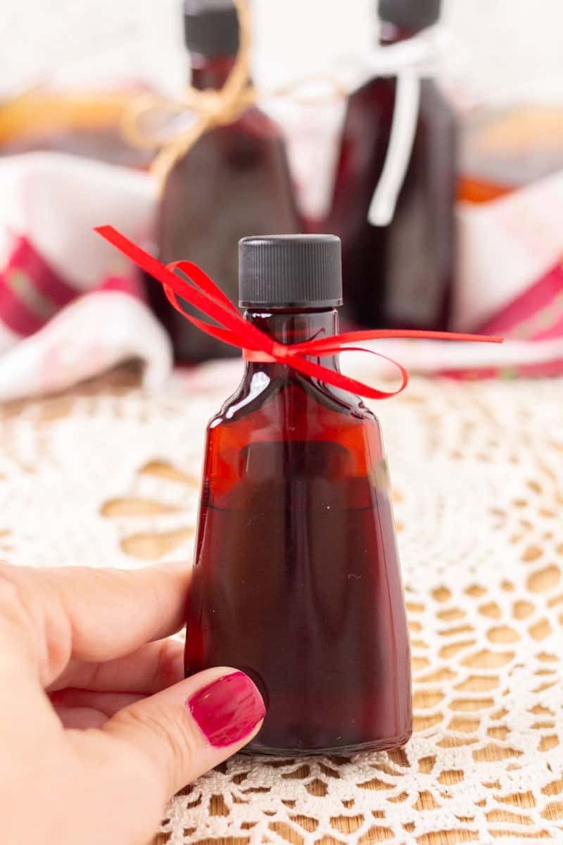 Easy Bourbon Vanilla Extract | thewendthousekitchen.com A hand holding a small amber bottle of vanilla extract with a red bow. In the background, a lace tablecloth and red and white towel.