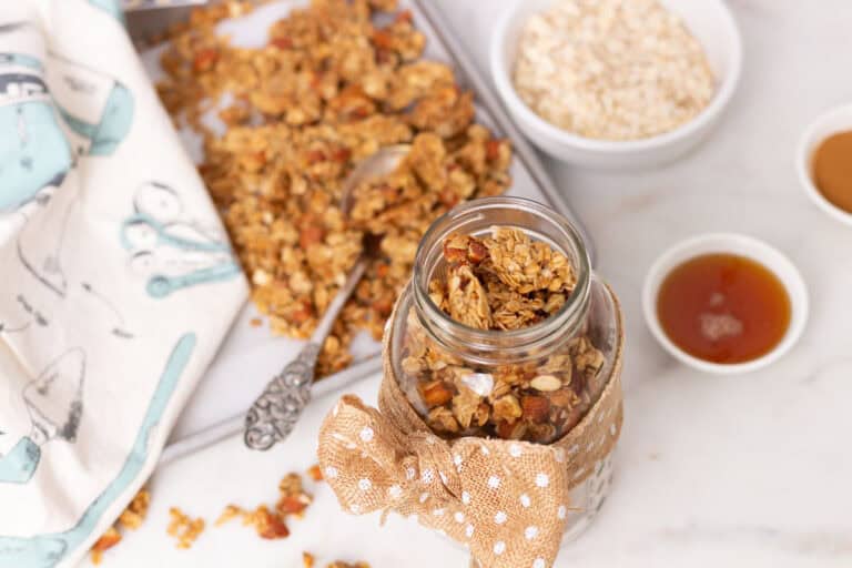 Homemade granola recipe in a Mason jar with some on a cookie sheet in the background. Ingredients in individual bowls around as well.