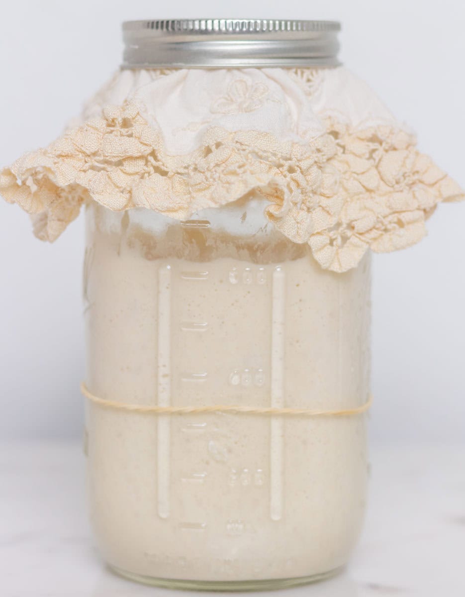 How To Make Sourdough Starter | thewendthousekitchen.com Sourdough starter in a Mason jar with a doily and a metal lid.