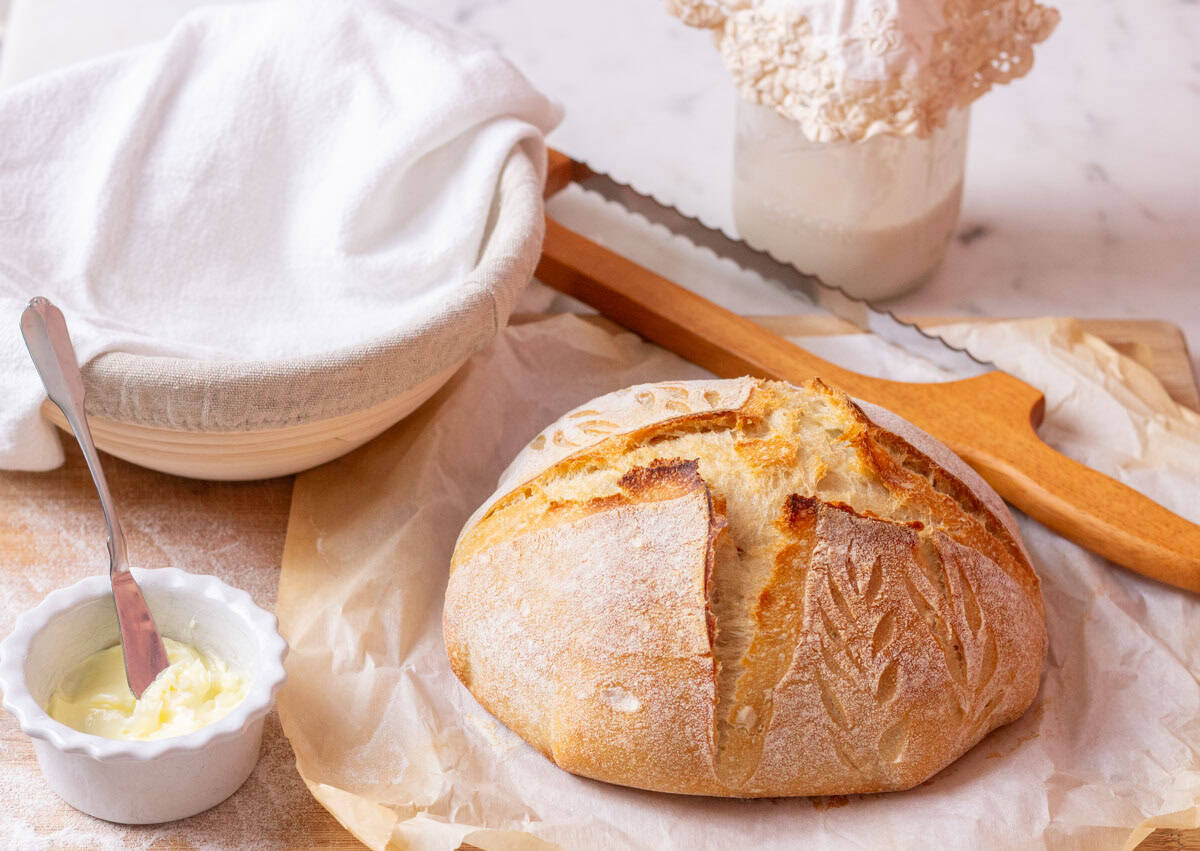 How To Make Artisan Sourdough Bread | thewendthousekitchen.com A loaf of baked sourdough bread with a bread knife in the background, a covered banneton and a jar of sourdough starter.