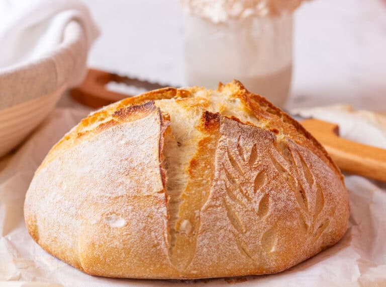 Baked sourdough bread on a tea towel with a bread knife in the background and a jar of sourdough starter