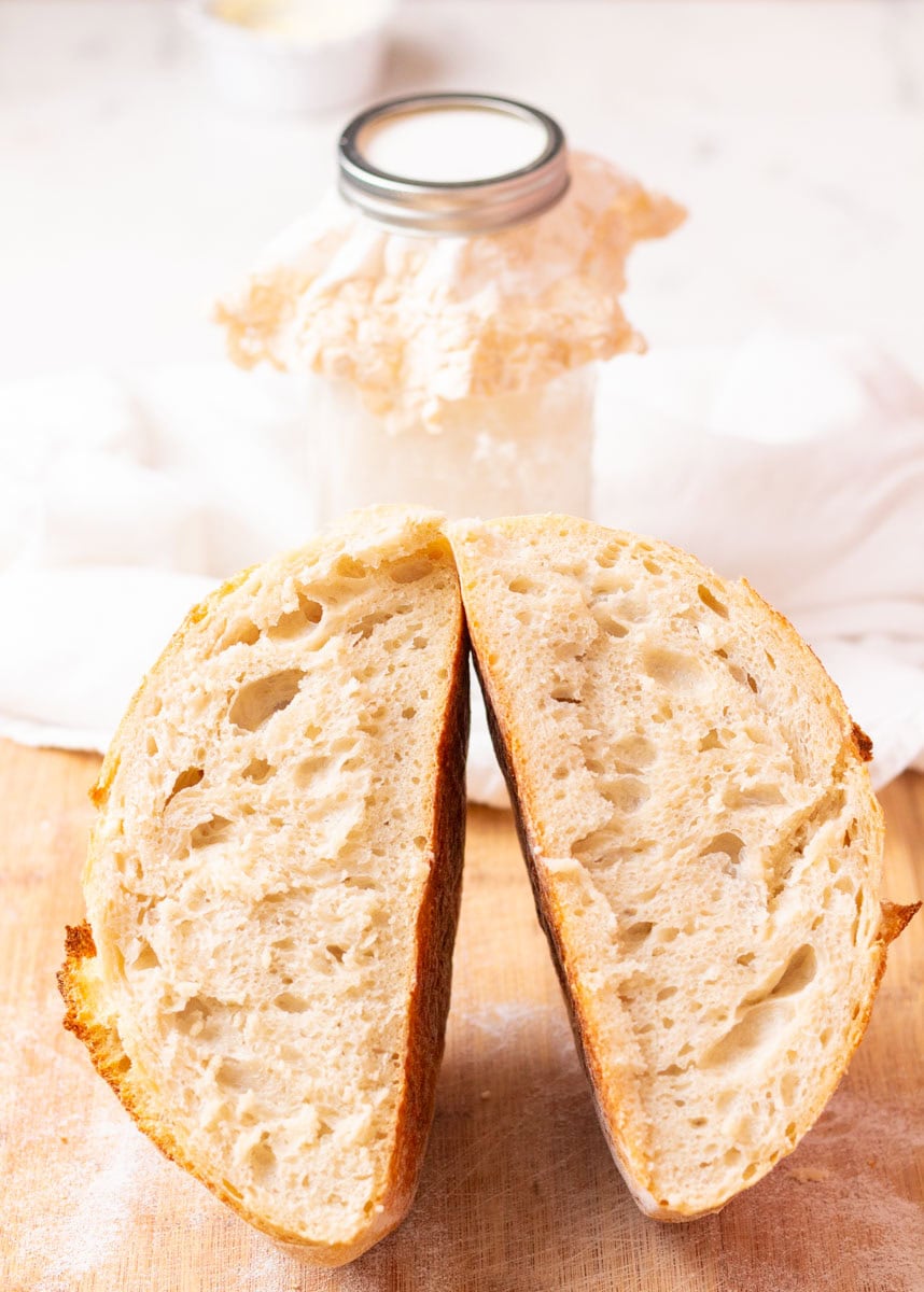 How To Make Artisan Sourdough Bread | thewendthousekitchen.com A baked loaf of sourdough bread cut in half with the insides showing and a jar of sourdough starter in the background.