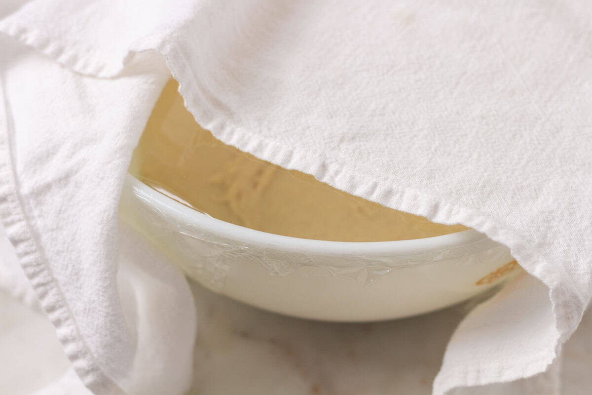 Sour Dough Discard Cinnamon Rolls | thewendthousekitchen.com Cinnamon roll dough in a large white Pyrex bowl covered with plastic wrap and partially covered with a white tea towel.