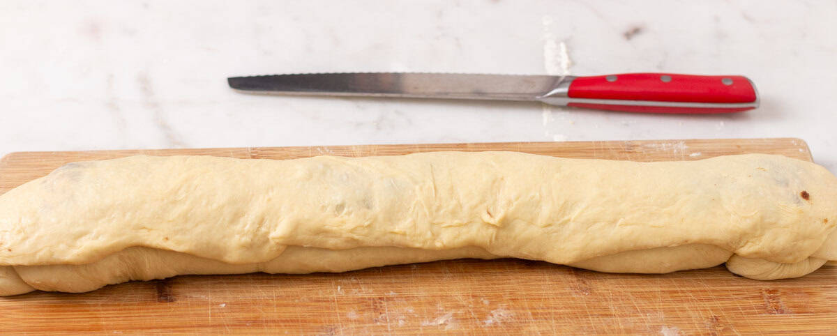 Sour Dough Discard Cinnamon Rolls | thewendthousekitchen.com Cinnamon roll dough rolled into a log on a wooden cutting board. A large, red serrated knife in the background.