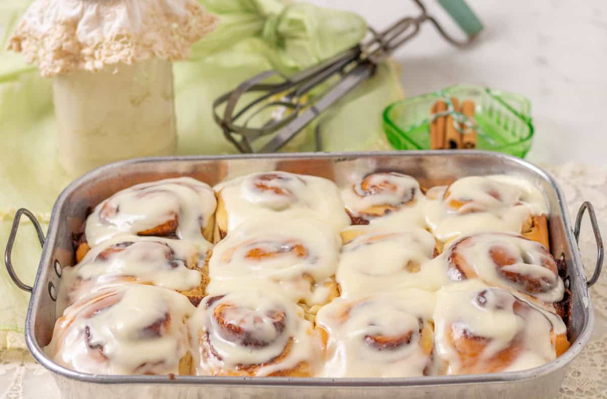 Iced cinnamon rolls in a metal pan with a jar of discard in the background and a manual mixer in the background.