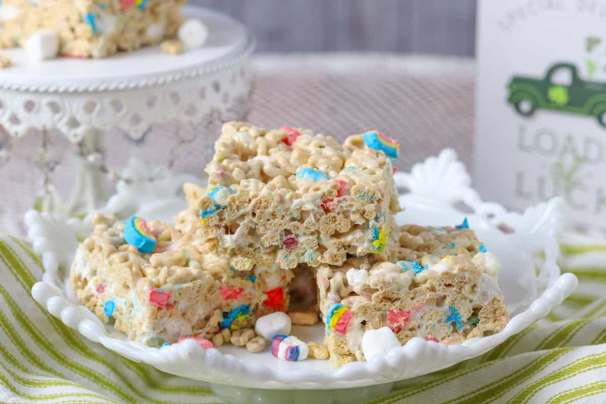 Lucky charm krispie treats on a white plate with more in the background on a white pedestal.