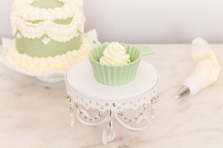 Italian meringue buttercream inside of a small green bowl with a decorated cake in the background and a piping bag.