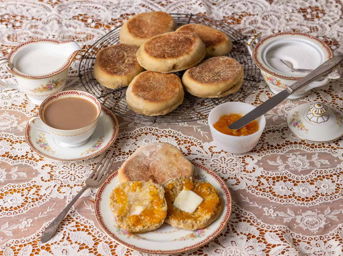 Sourdough Discard English Muffin Recipe | thewendthousekitchen.com Overhead table shot on a lace tablecloth with English muffins on a cooling grid, a cup of coffee, a cup of jam, a cream and sugar bowl and an English muffin on pretty china with butter and jam.