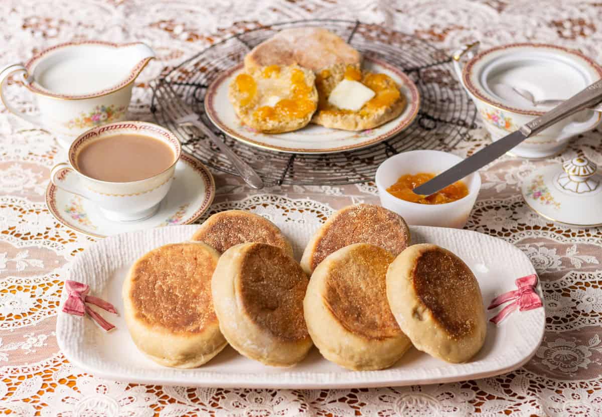 Sourdough Discard English Muffin Recipe | thewendthousekitchen.com Sourdough discard English muffins on a plate with a cup of coffee in the background, a sugar and creamer bowl and buttered English muffins with jam on a plate.