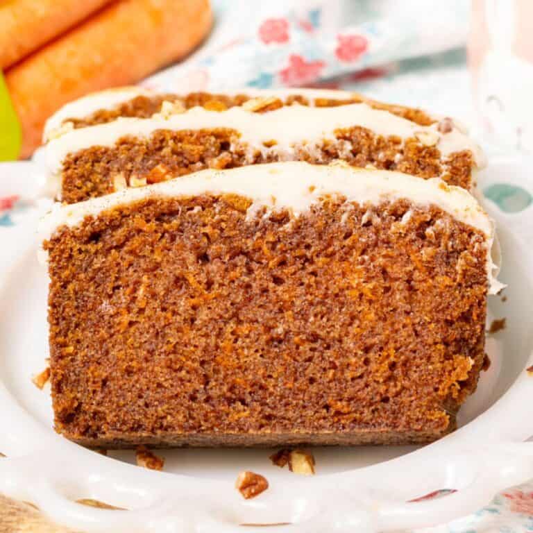 Slices of carrot cake loaf with cream cheese frosting on the top and carrots in the background.