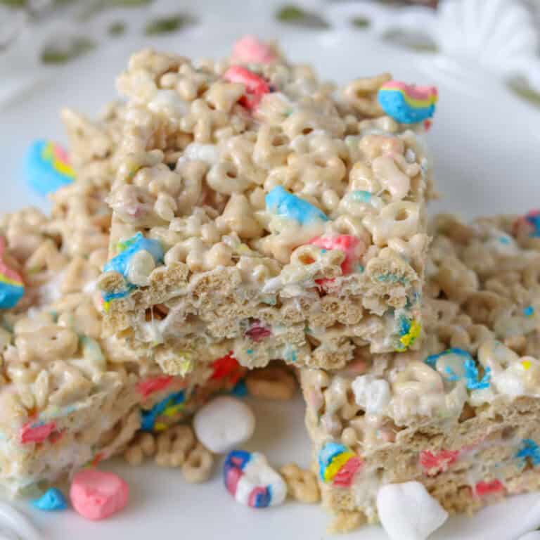 Lucky Charm krispie treats stacked on a white plate with marshmallows and cereal scattered around.
