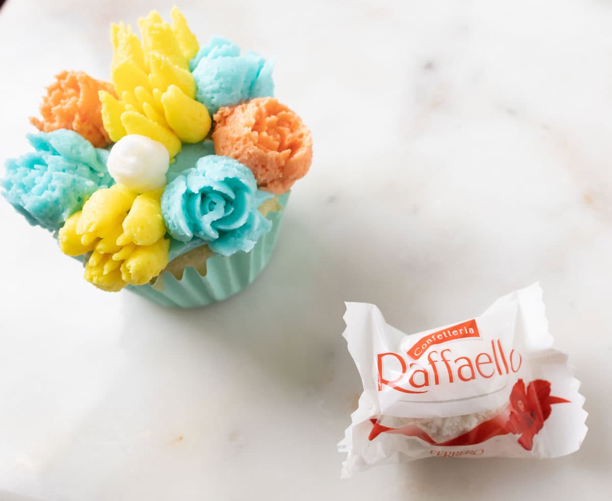 Bunny Cupcakes for Easter (Bunny Butt Cupcakes) | thewendthousekitchen.com A photo of a cupcake decorated with turquoise, yellow and orange buttercream flowers and a large white buttercream dot in the middle. A wrapped Raffaello candy next to the cupcake.