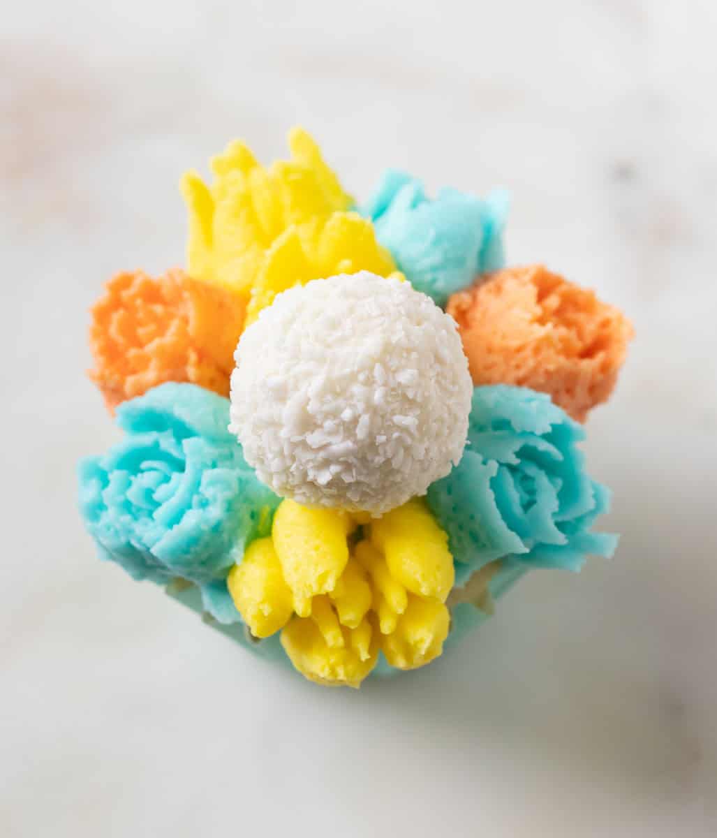 Bunny Cupcakes for Easter (Bunny Butt Cupcakes) | thewendthousekitchen.com A cupcake decorated with turquoise, orange and yellow buttercream flowers and a Raffaello candy in the middle of the cupcake.