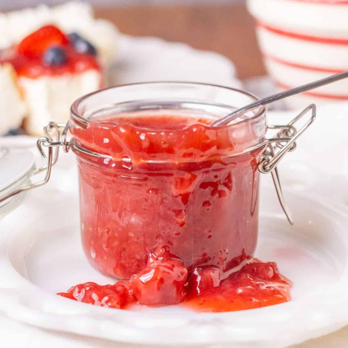 Strawberry sauce in a clear jar with a spoon on a white plate with strawberry sauce falling down the sides.