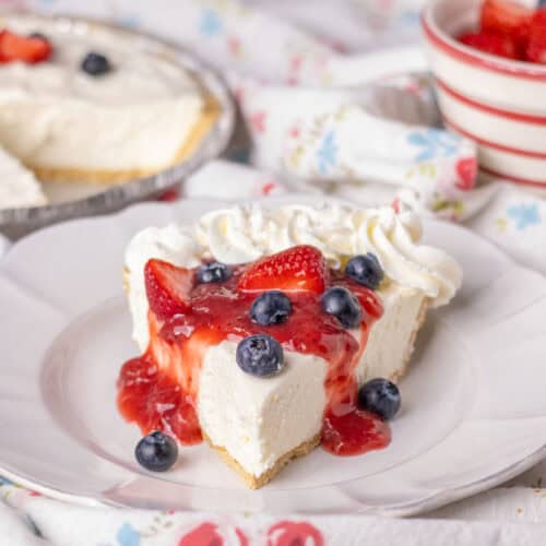 A slice of no bake cheesecake with strawberry sauce and fresh strawberries and blueberries. The rest of the pie in the background.