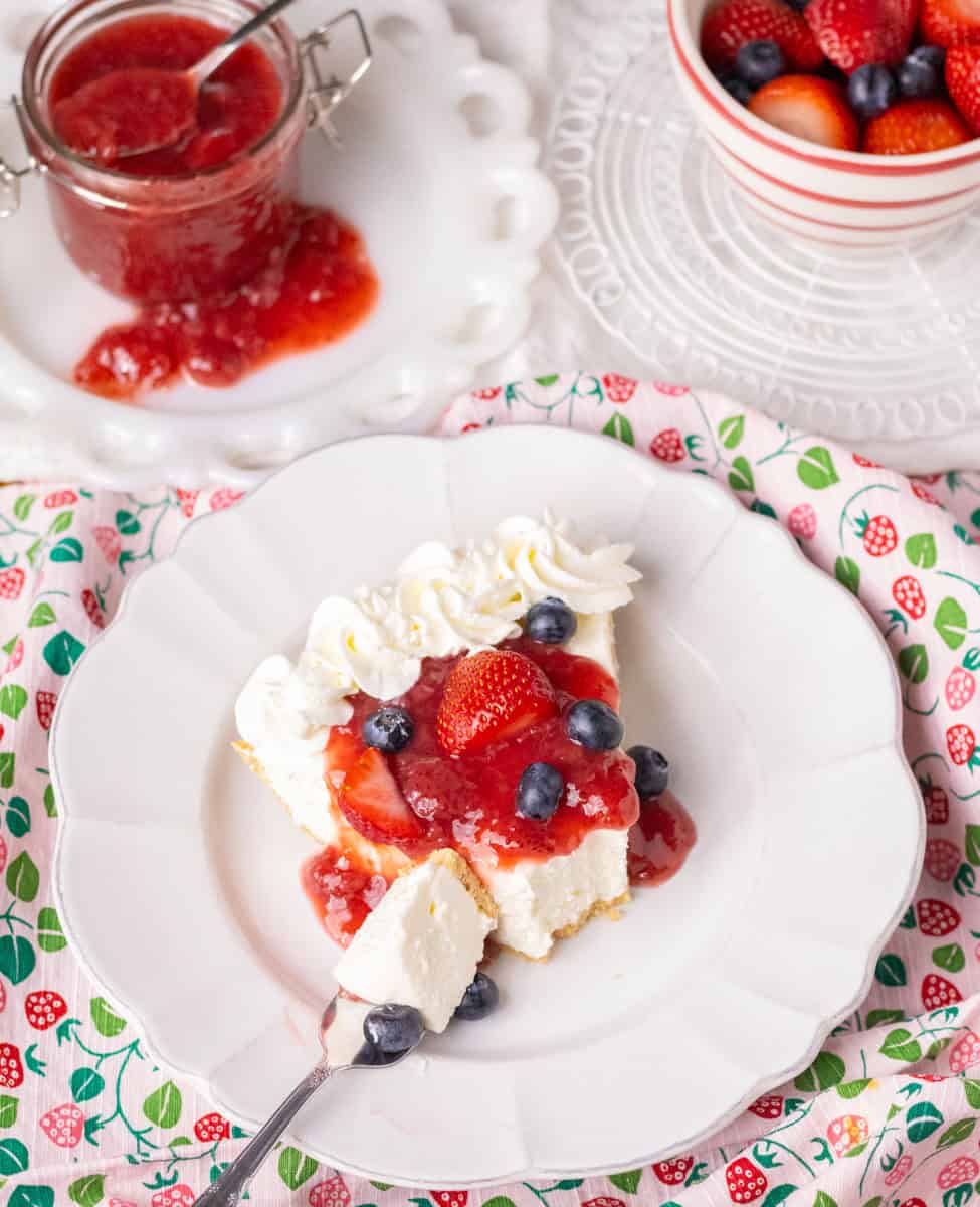 3 Ingredient No Bake Cheesecake | thewendthousekitchen.com Piece of no bake cheesecake on a white plate with a forkful taken out. Has strawberry sauce and fresh berries. On a pink dish towel with strawberries.