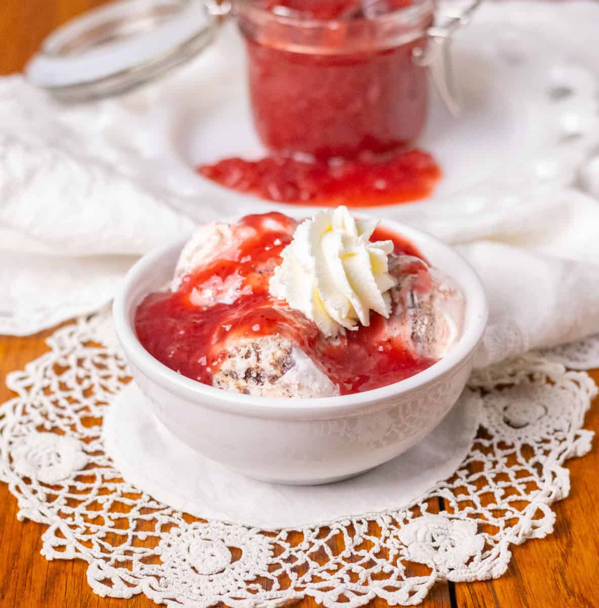 How To Make Strawberry Sauce | thewendthousekitchen.com Strawberry sauce on a small bowl of ice cream with whipped cream. Jar of strawberry sauce in the background on a white plate.