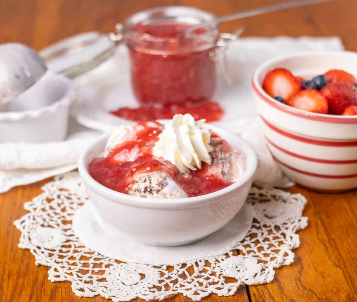 How To Make Strawberry Sauce | thewendthousekitchen.com A photo of ice cream in a small white bowl with strawberry sauce and whipped cream on top. Berries in the background and a glass jar with strawberry sauce.