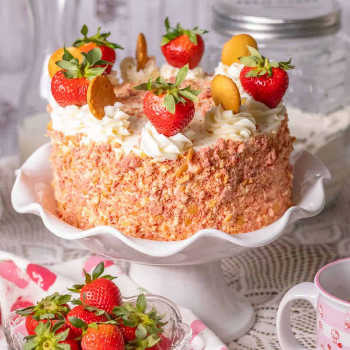Strawberry crunch cake on a white pedestal cake plate with strawberries in a bowl in the front.