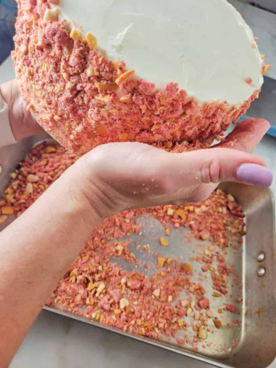 Strawberry Crunch Cake | thewendthousekitchen.com Holding a frosted cake with white buttercream and pressing strawberry crunchies onto the side of the cake. A roasting pan is catching the strawberry crunchies.