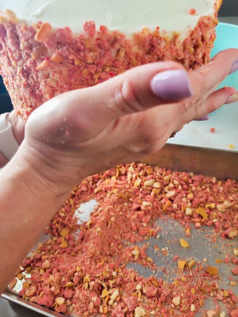 Strawberry Crunch Cake | thewendthousekitchen.com Holding an 8-inch frosted cake with white buttercream and patting strawberry crunchies onto the side of the cake. The roasting pan is catching the strawberry crunchies as they fall.