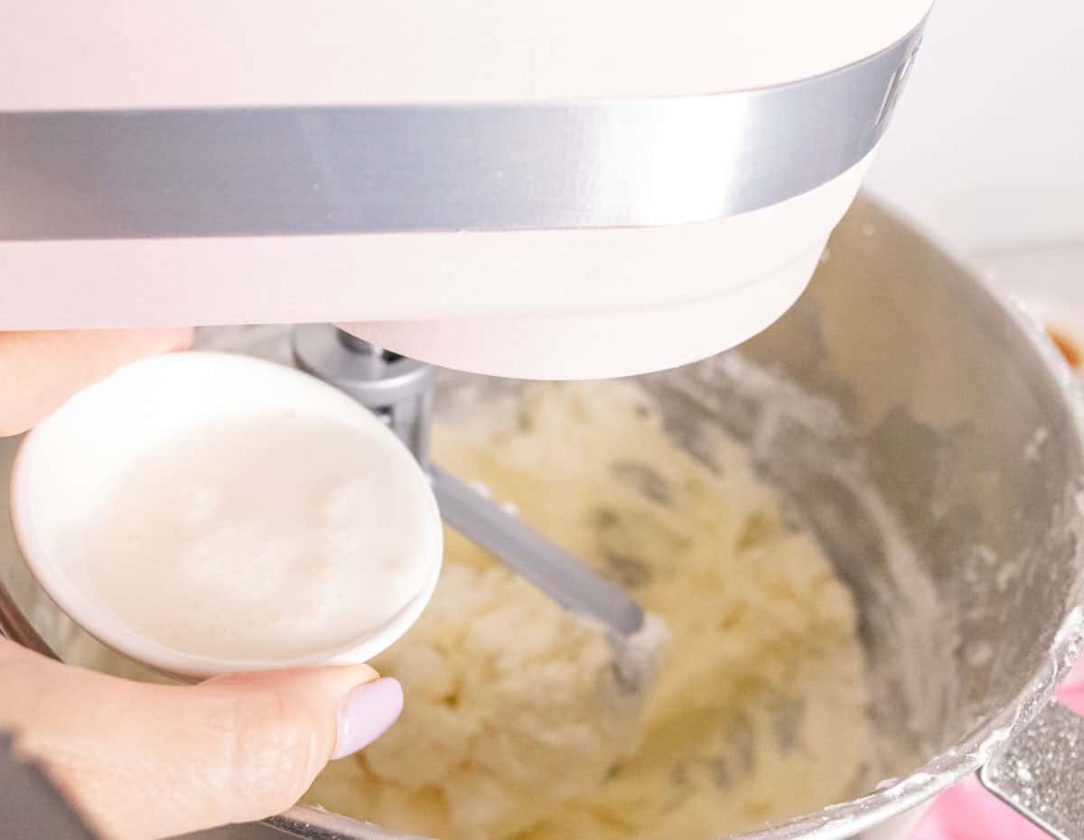 Strawberry Crunch Cake | thewendthousekitchen.com Pouring whipping cream into the buttercream in the bowl of a stand mixer.