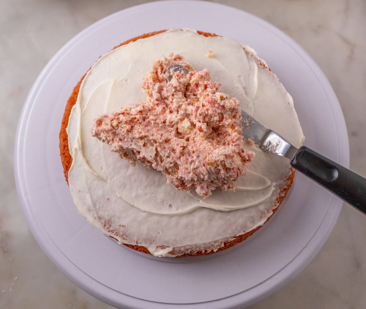 Strawberry Crunch Cake | thewendthousekitchen.com Spreading buttercream and strawberry crunchy mixture on a lightly frosted cake layer with a small offset spatula.